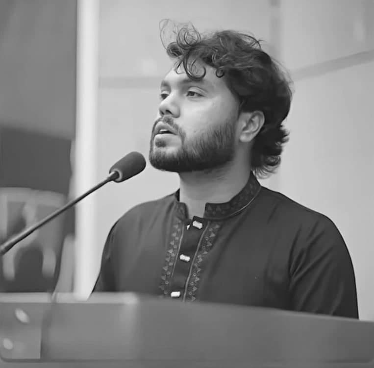 Sharif Osman bin Hadi in a dark panjabi, speaking at a lectern microphone.